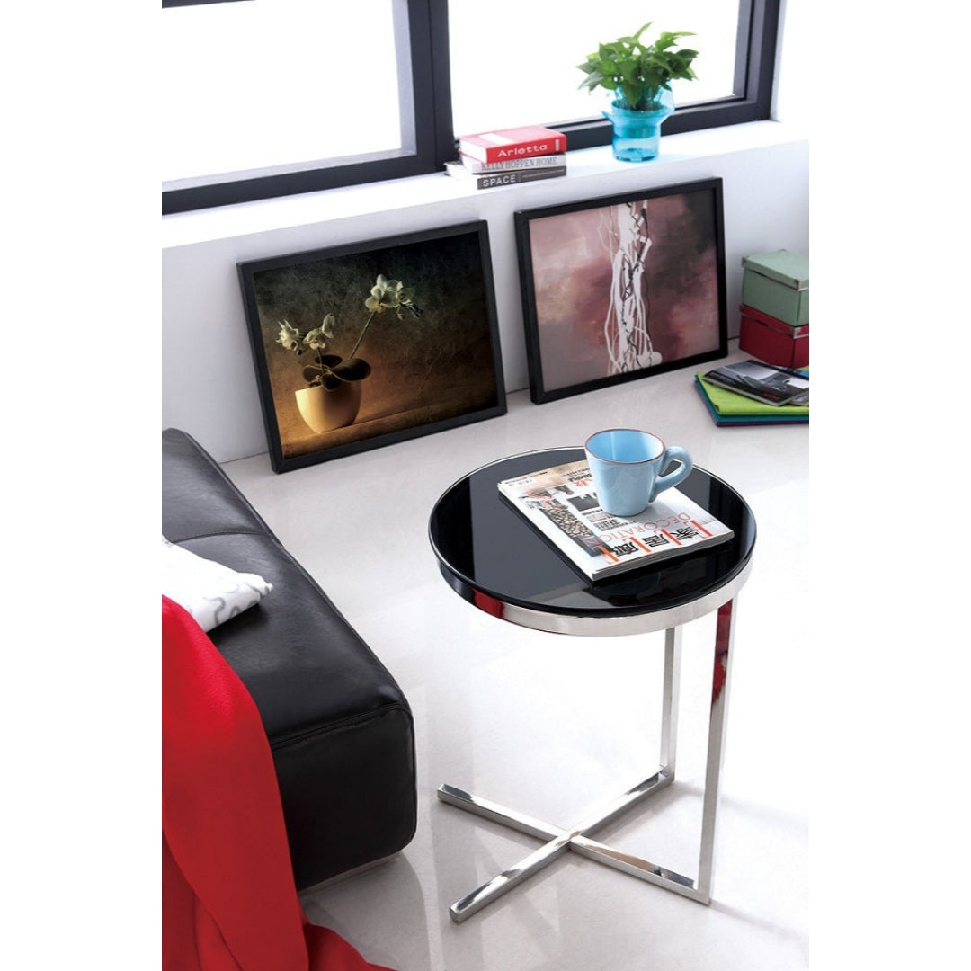 A polished stainless steel side table with black tempered glass top, placed in a room next to a red chair with a magazine and a cup on top of it, featuring mirror polished finish.