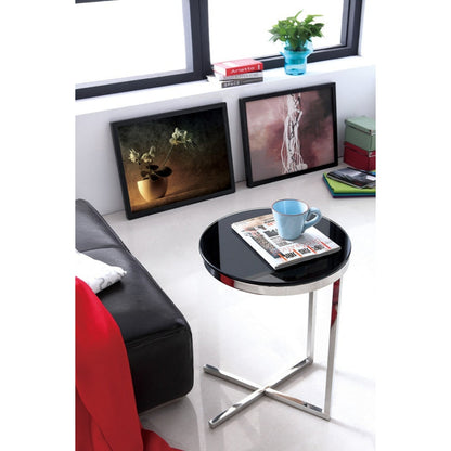 A polished stainless steel side table with black tempered glass top, placed in a room next to a red chair with a magazine and a cup on top of it, featuring mirror polished finish.