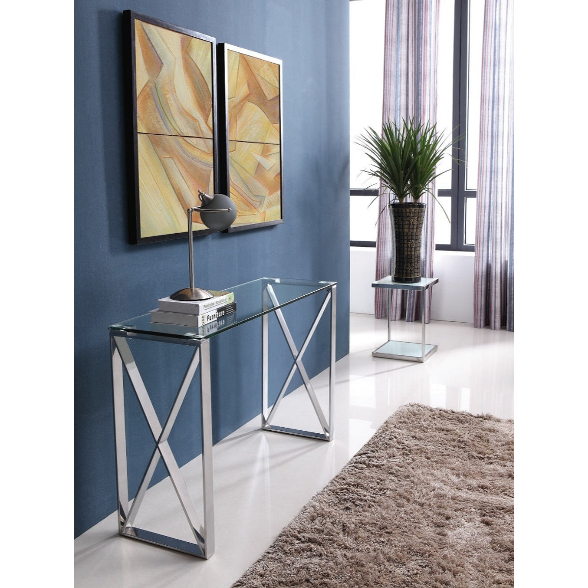 A polished stainless steel console table with a clear glass top, placed against a blue wall with a framed abstract painting above it. A potted plant and a decorative lamp are on the console, set on a white carpet.