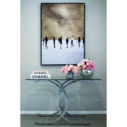 A polished stainless steel console table with a clear glass top, displayed in a room with a decorative mirror and a painting above it. A Chanel brand sign and a vase with pink flowers are also visible on the table.